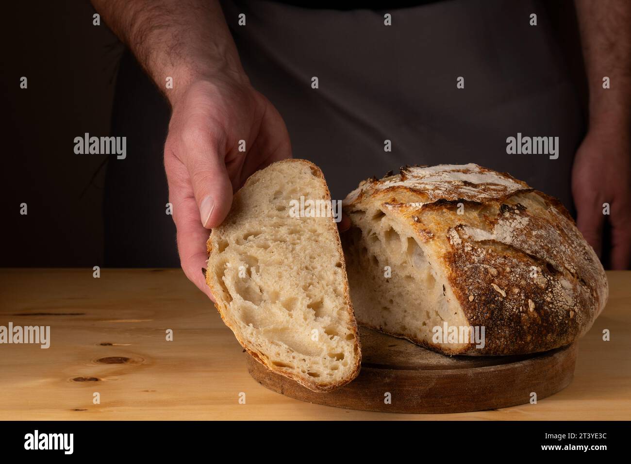 The hands of a young man handling sourdough bread, highlighting the bread with beautiful golden tones against the dark background. Stock Photo