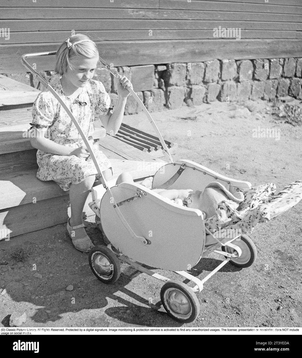 Summer in the 1940s. A girl is sitting on the stairs rocking a pram where her little sibling is lying. She is wearing a summer dress to be nice, but her shoes are very worn. Sweden 1947. Kristoffersson ref V93-5 Stock Photo
