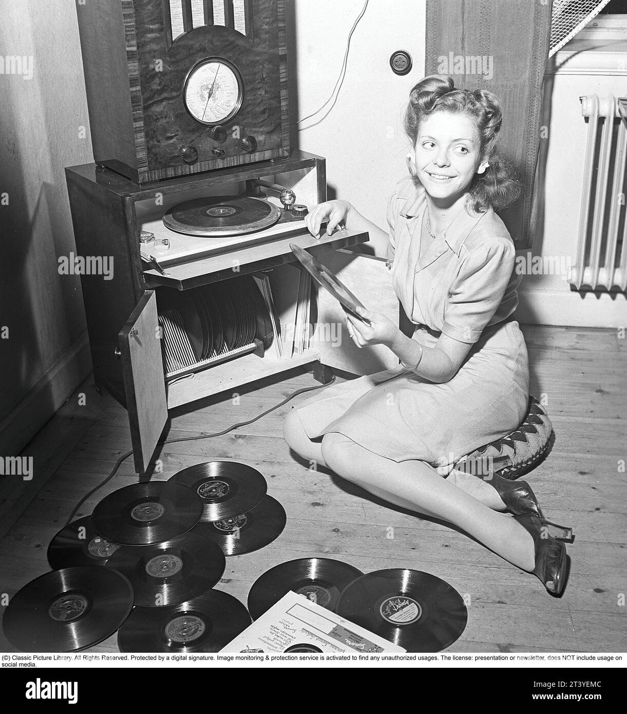 Woman at a gramophone in the 1940s. She has lots of different records to choose from to listen to. A radio is on top of the gramophone.  The records were made of fragile material and the speed of the record to sound as it should was 78 revolutions per minute. Note the small tin boxes beside the gramophone. They contain the needles that are the medium to read the records with, the needle running in the fine lines of the record. The needles needed to be replaced as they became worn, and you did this yourself.    Sweden 1945. Kristoffersson ref O124-5 Stock Photo