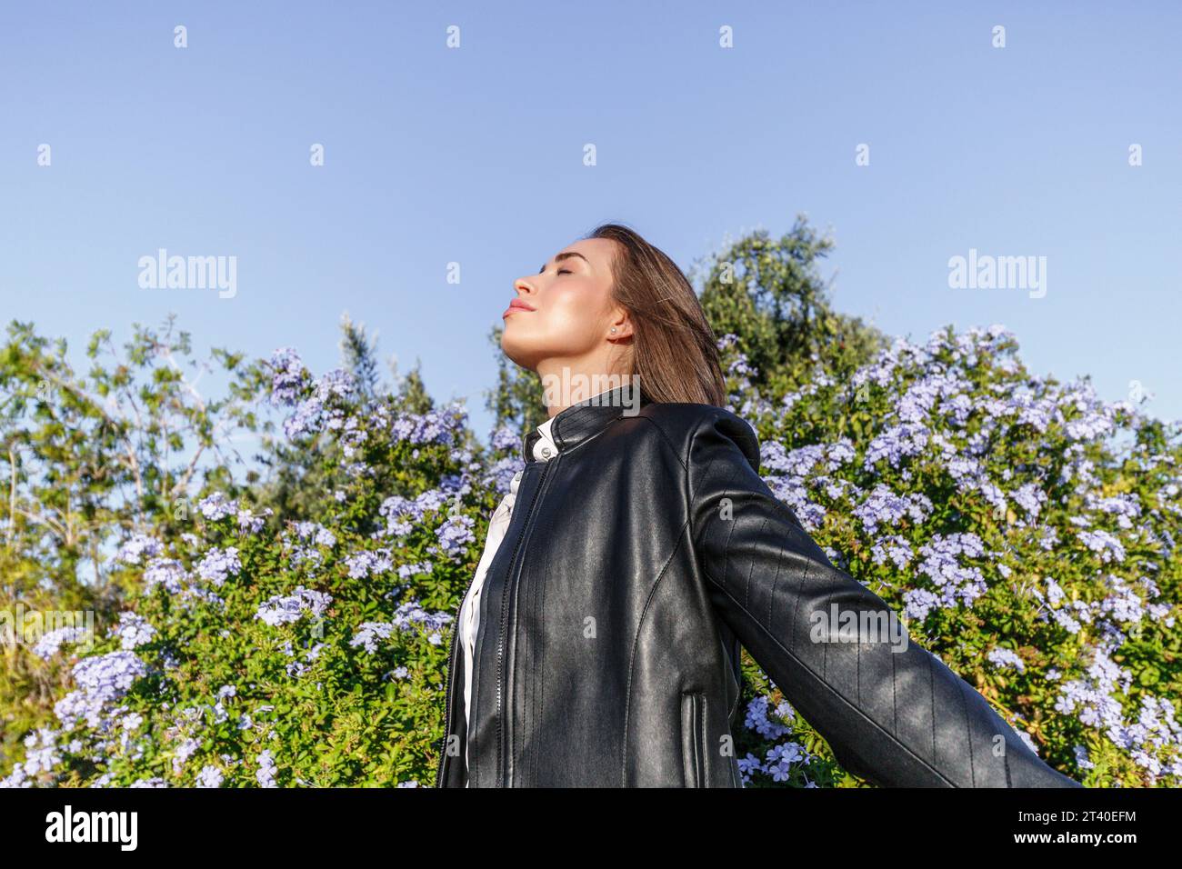 pretty young woman with blonde hair and dressed in a leather jacket, with her eyes closed breathing fresh air in the forest on a sunny day Stock Photo