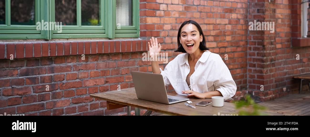 Friendly asian woman sitting with laptop, waving at you, saying hello ...