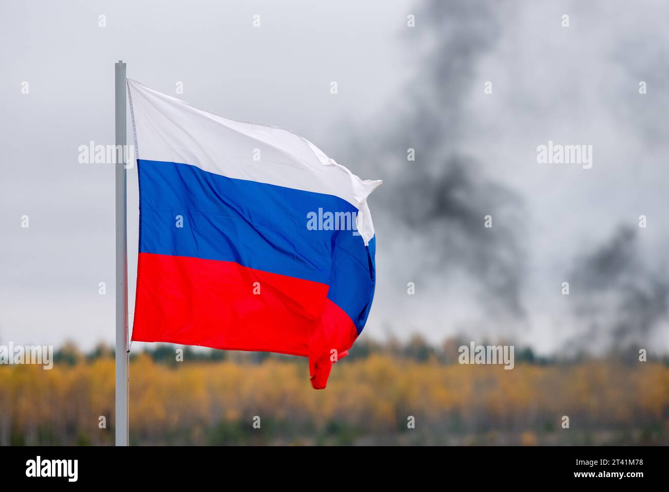 The white-blue-red flag of the Russian Federation is flying in the wind against the background of black smoke in a cloudy sky. Stock Photo