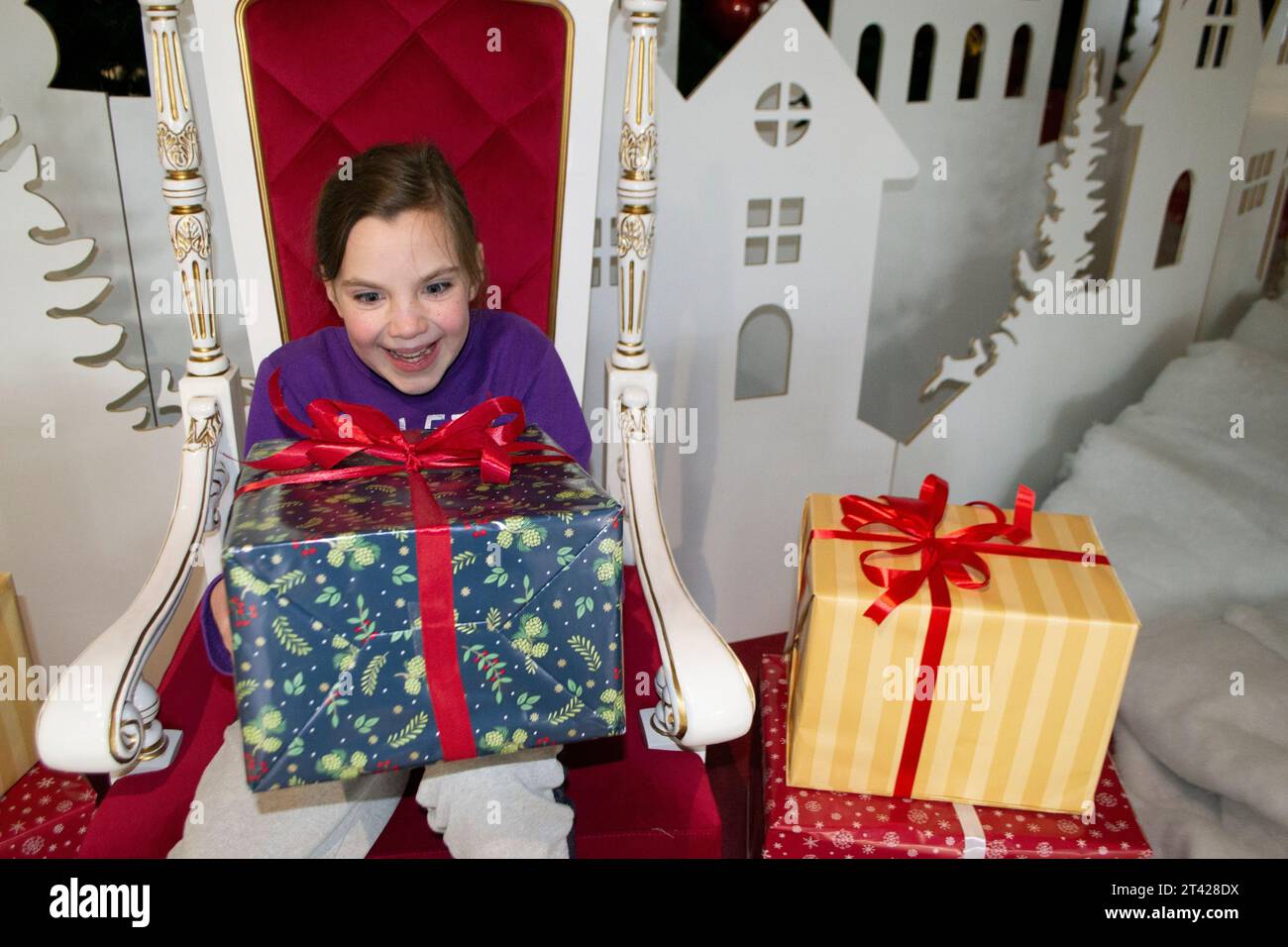 Happy little girl in Santa's hat sitting in a room decorated for Christmas. Portrait of adorable girl playing with Christmas gifts near New Year tree. Stock Photo