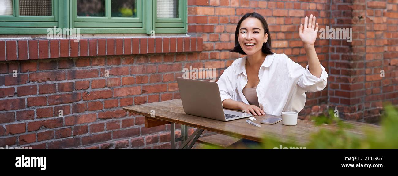 Friendly asian woman sitting with laptop, waving at you, saying hello ...