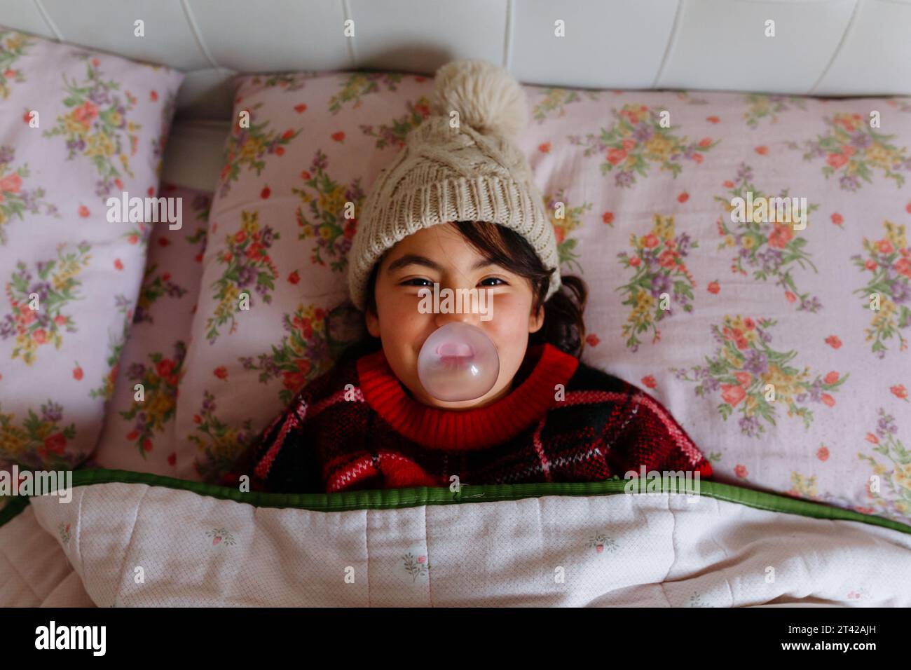 Girl wearing beige warm hat making bubble lying in the bed under