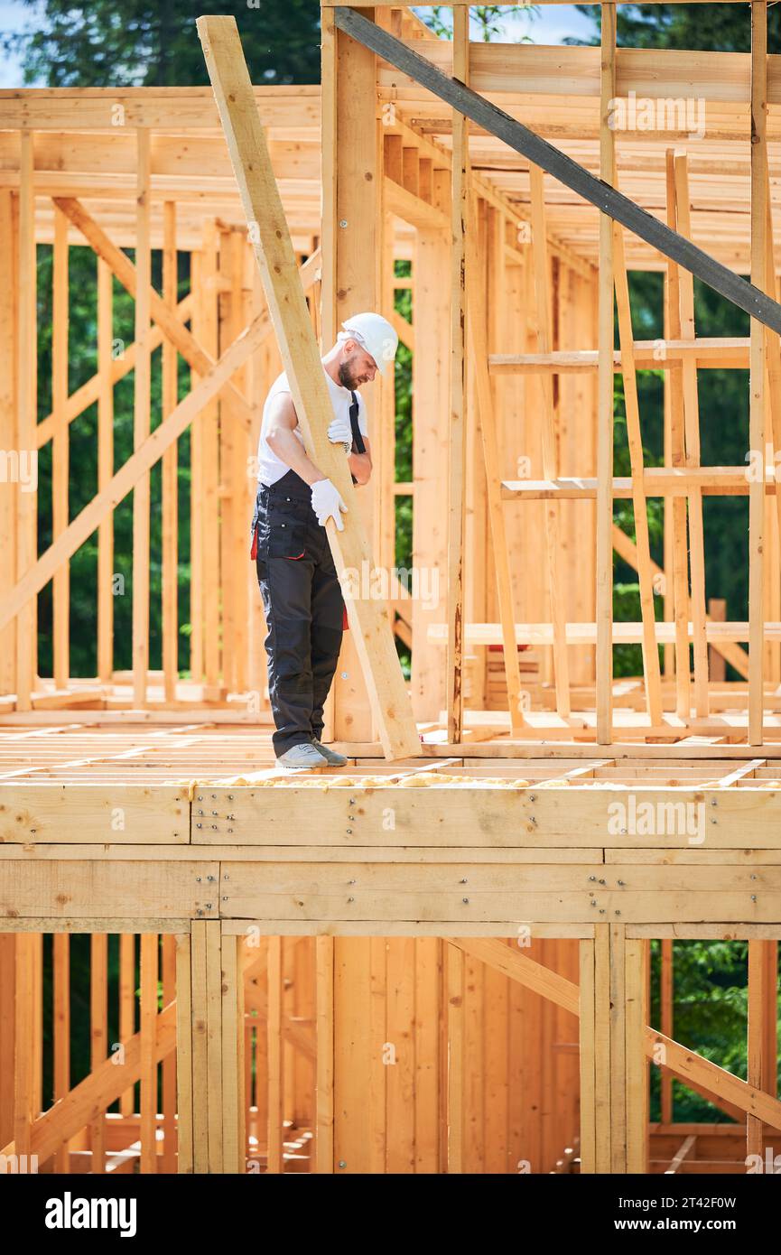 Carpenter constructs wooden-framed house. Man holds large beam in his hands while wearing work clothes and helmet. Idea of modern, eco-friendly construction. Stock Photo