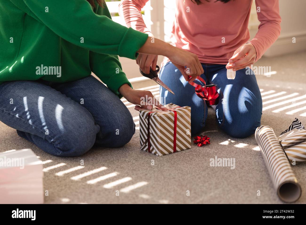 Diverse mother and daughter kneeling on floor, decorating present at home, copy space Stock Photo