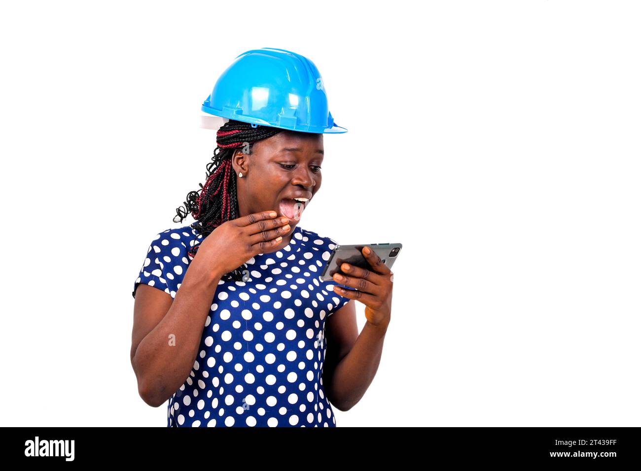 young female engineer wearing blue safety helmet surprised and watching video on mobile phone while smiling. Stock Photo