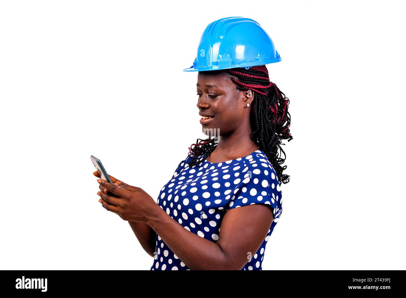 a beautiful female engineer with safety helmet standing on white background reading message on cellphone, smiling. Stock Photo