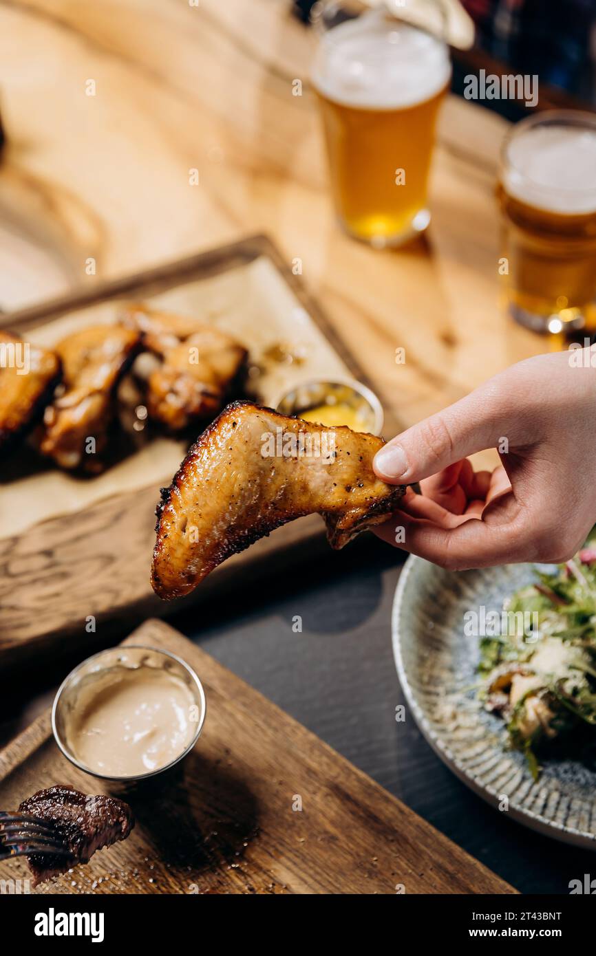 Male hand holding spicy grilled BBQ chicken wings and eating with beer in pub Stock Photo