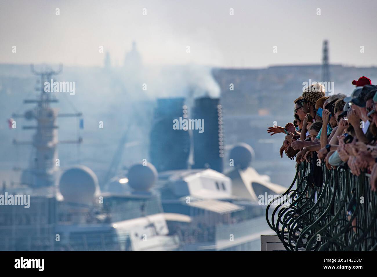 Thick smoke spues from the chimneys of a vast cruise ship moored in Malta's capital, Valletta whilst large numbers of tourists watch an event. Stock Photo