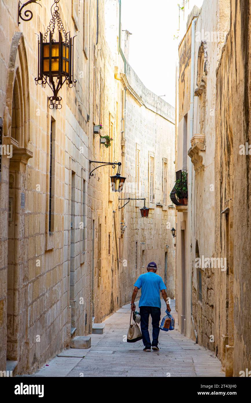 A man walks doen a narrow alleyway with his shopping in Mdina, Malta Stock Photo