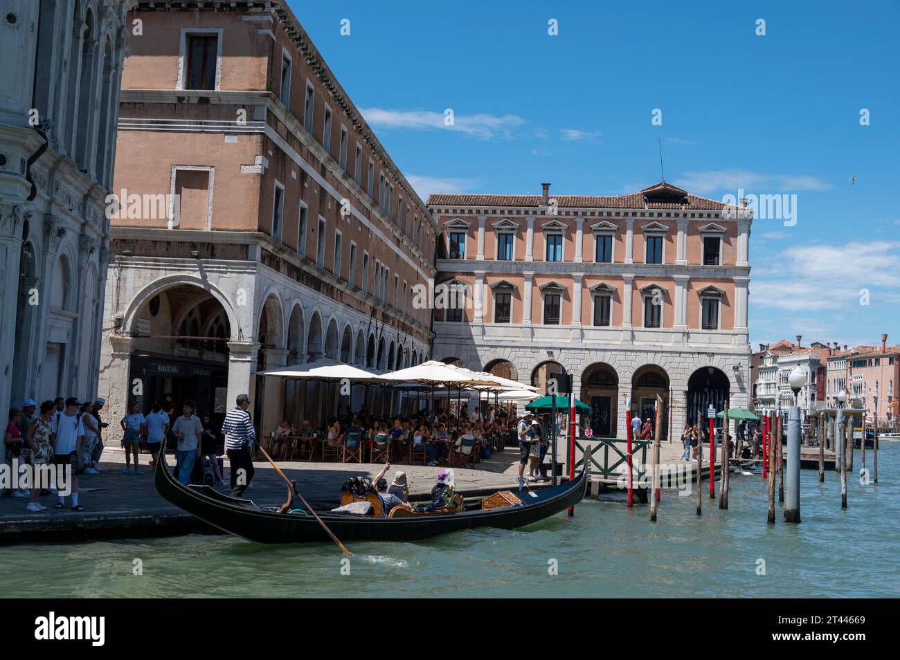 A gondola passing a small square with an outdoor café and near the Rialto fresh fruit and veg market on the banks of the Grand Canal in Venice in the Stock Photo