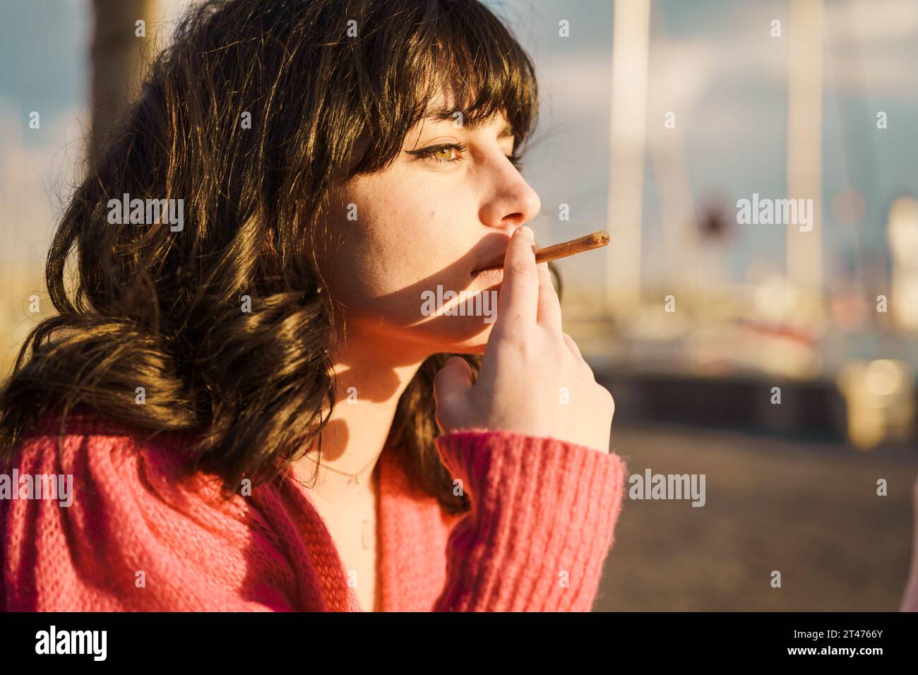 Young woman smoking by the sea - A young woman smokes by the sea, reflecting on youth's vices. The calm setting contrasts with her intense gaze, highl Stock Photo