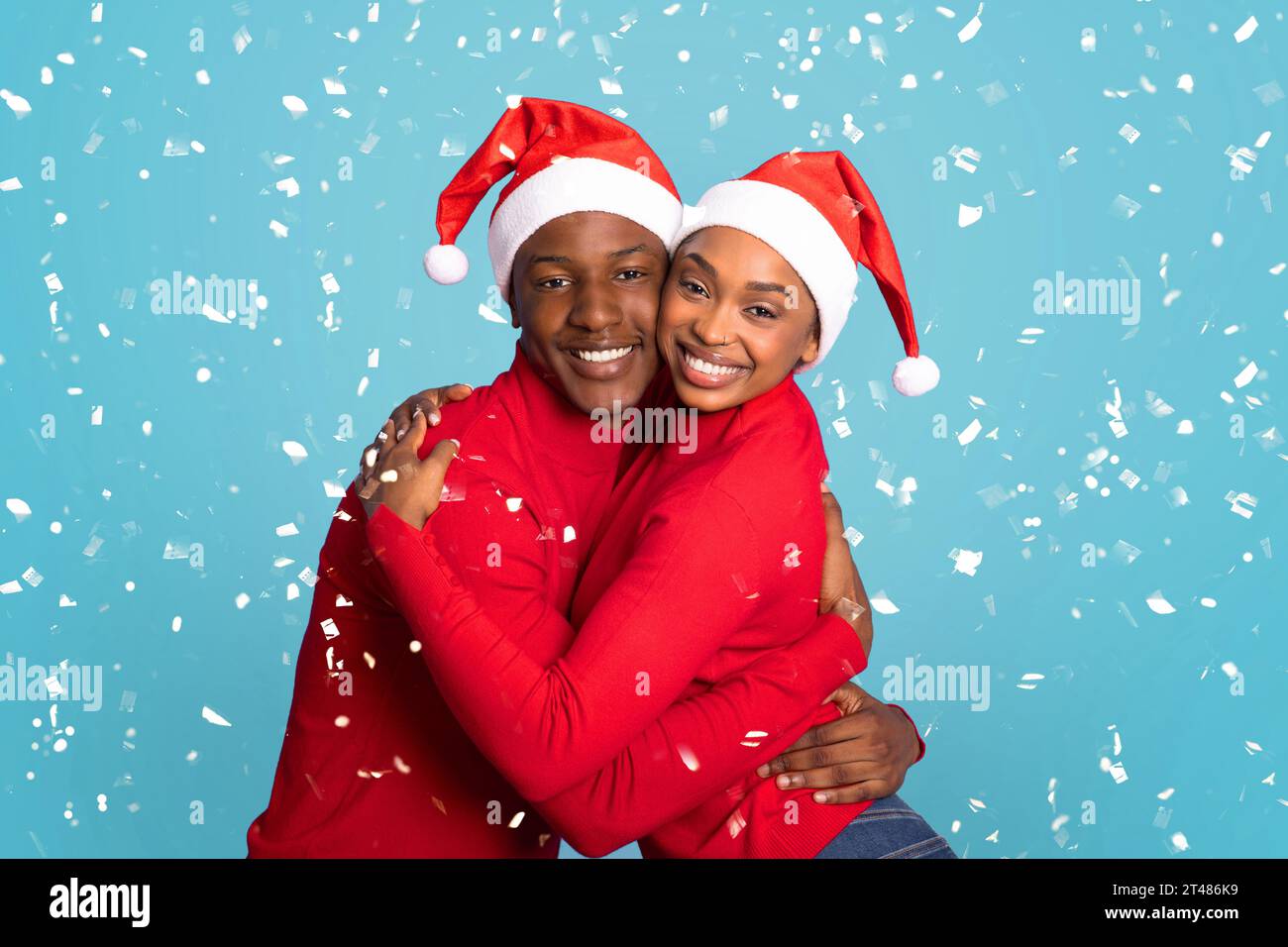Black Couple In Santa Hats Hugging Under Falling Confetti, Studio Stock Photo