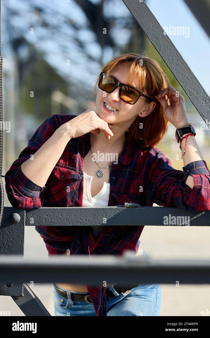 A fashionable redhead woman enjoying an autumn evening in the city. She's wearing a trendy red jacket, a white t-shirt, and denim jeans, making a bold Stock Photo