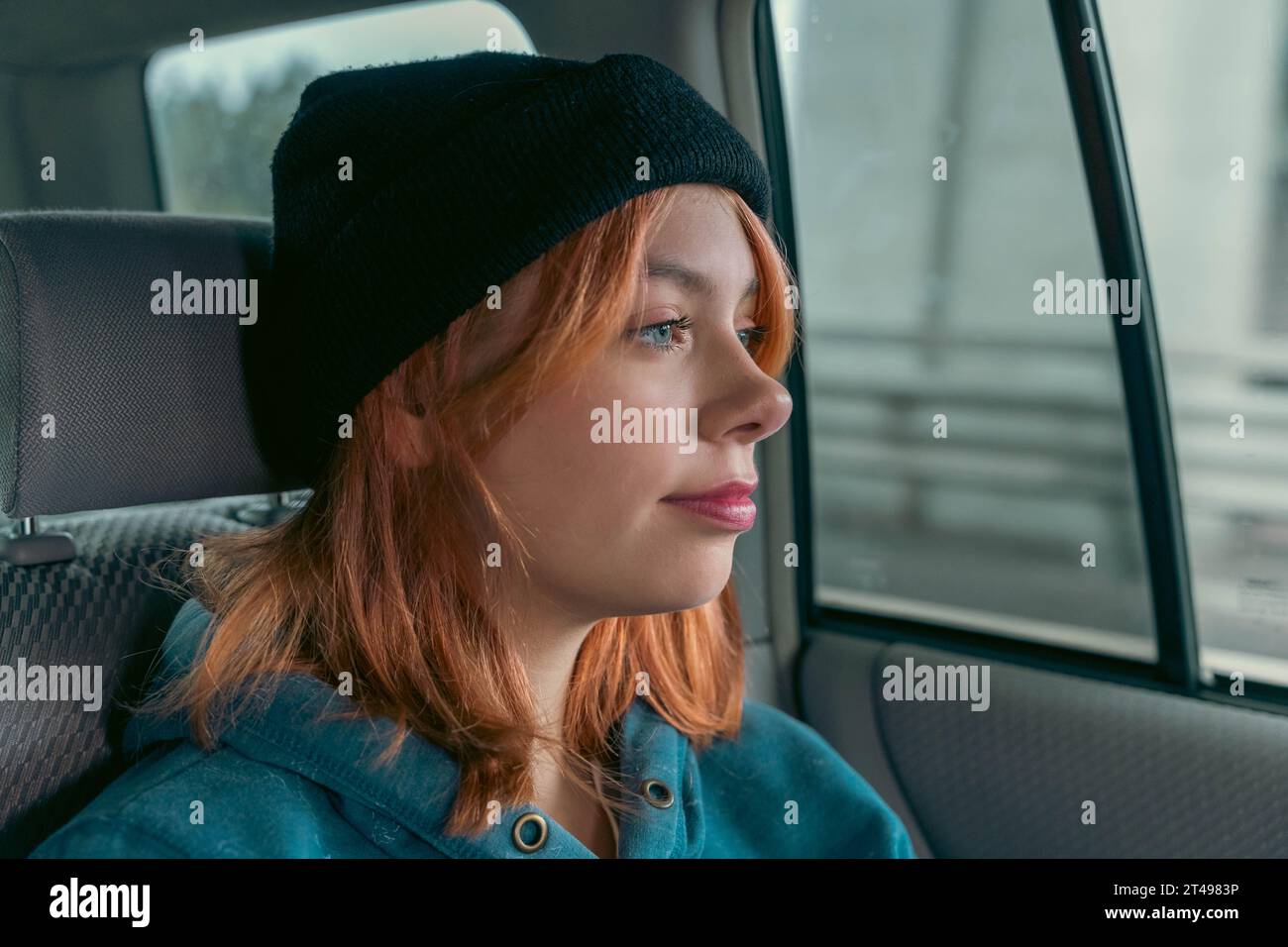 A beautiful girl in a black cap with pink hair relaxed in the back seat of the car, immersed in her thoughts. looking out the window, she is immersed Stock Photo
