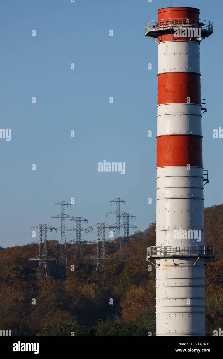 Red-and-white power plant pipe against the background of blue sky and forest power lines Stock Photo