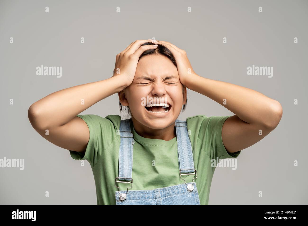 Desperated teen girl shouting holding head feeling intensive negative emotions on gray background.  Stock Photo