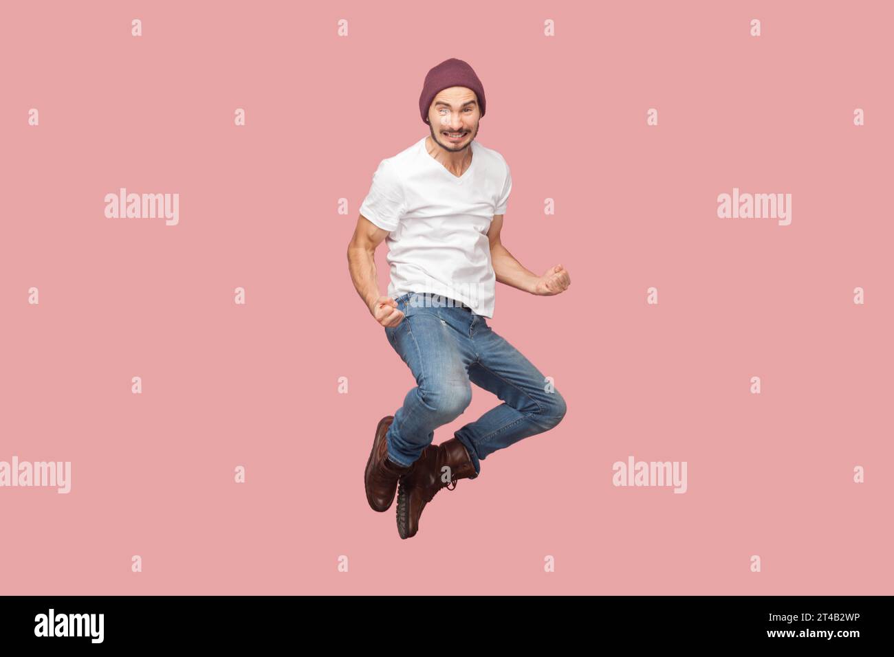 Portrait of extremely happy joyful bearded man in white T-shirt and beany hat jumping with clenched fists, rejoicing, celebrating victory. Indoor studio shot isolated on pink background. Stock Photo