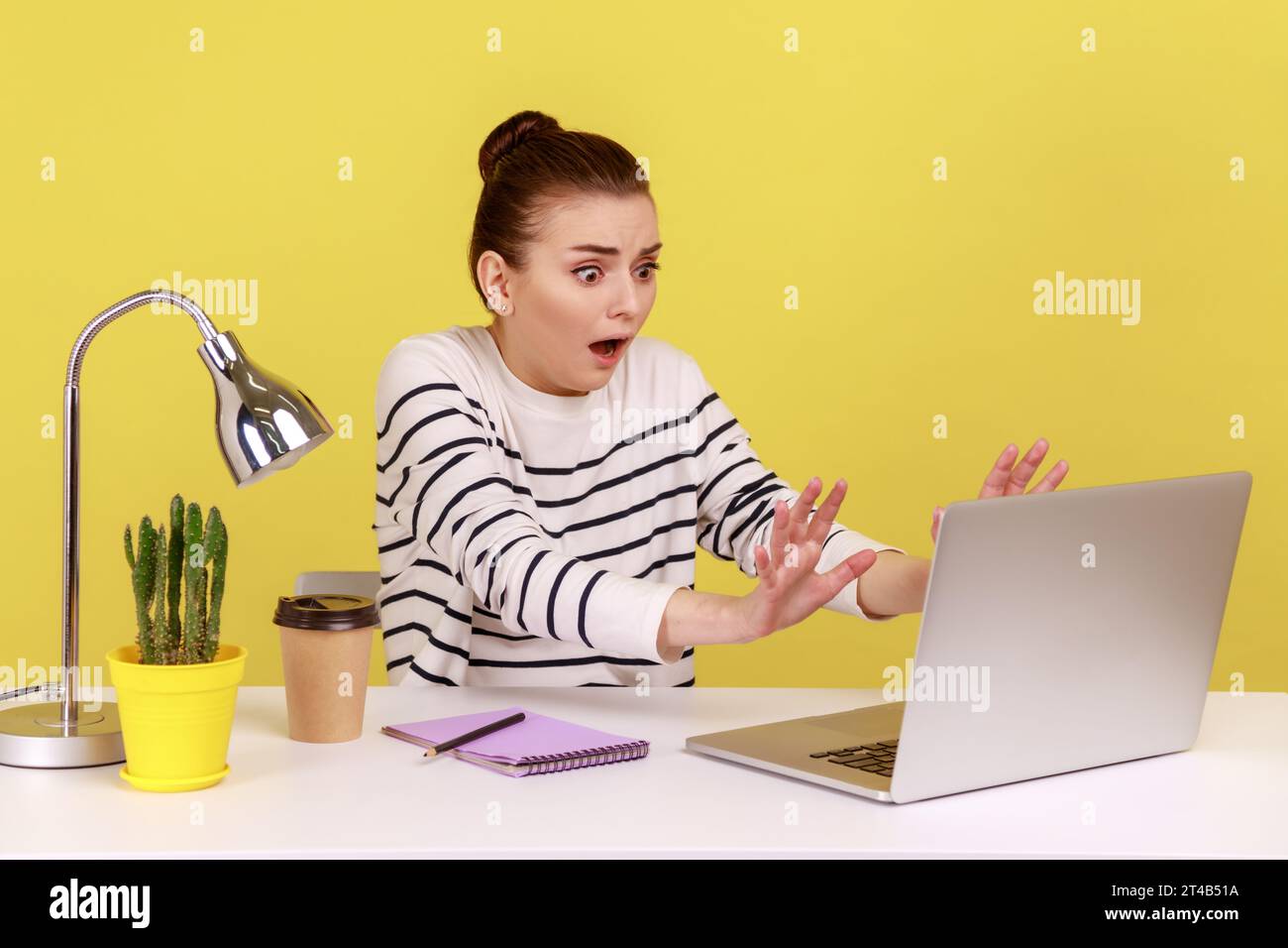Frightened woman with shocked expression raising hands in stop gesture, defending herself, working on laptop, sees prohibited content. Indoor studio studio shot isolated on yellow background. Stock Photo