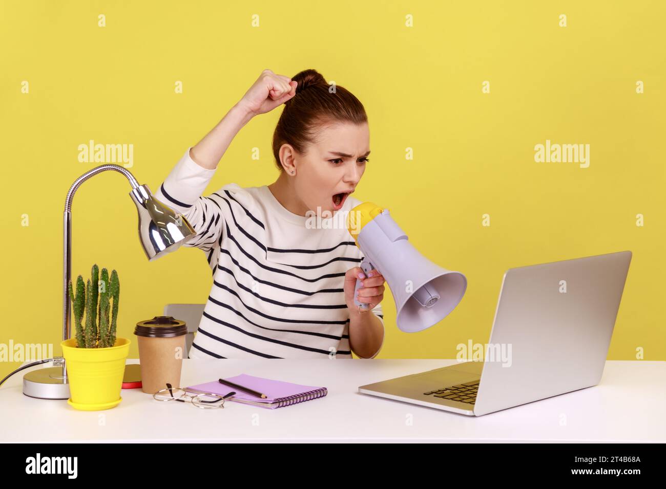 Portrait of angry woman loudly screaming at megaphone, making announce, protesting, sitting on workplace and looking at laptop screen. Indoor studio studio shot isolated on yellow background. Stock Photo