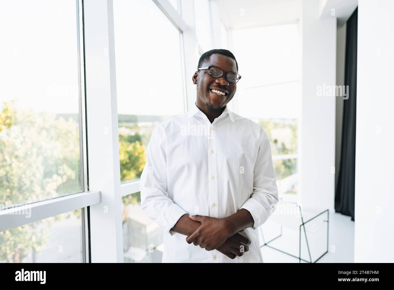 Young african man in white shirt and glasses posing near large window ...