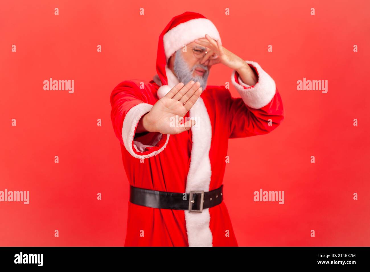 Santa claus in red costume pinching nose with fingers to avoid bad smell and showing stop gesture, demonstrating repulsion to stink, awful odor. Indoor studio shot isolated on red background. Stock Photo