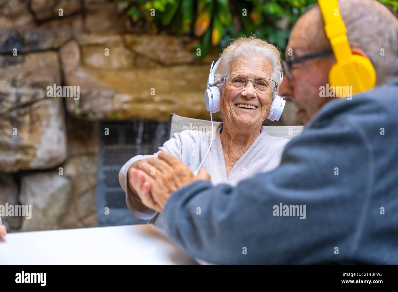 Happy elder people using headphones and smiling sitting in a garden in a geriatric Stock Photo
