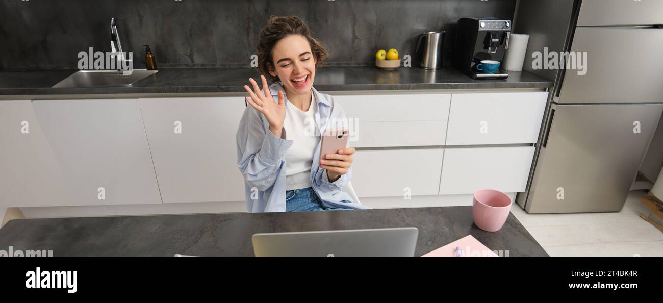 Chatty brunette woman sits at home, video chats with friend, waves hand at camera, holds smartphone and uses laptop Stock Photo