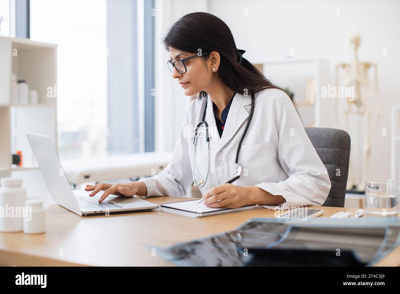 Side view of indian female doctor using portable computer while sitting at office desk in health center interior. Efficient mature general practitione Stock Photo