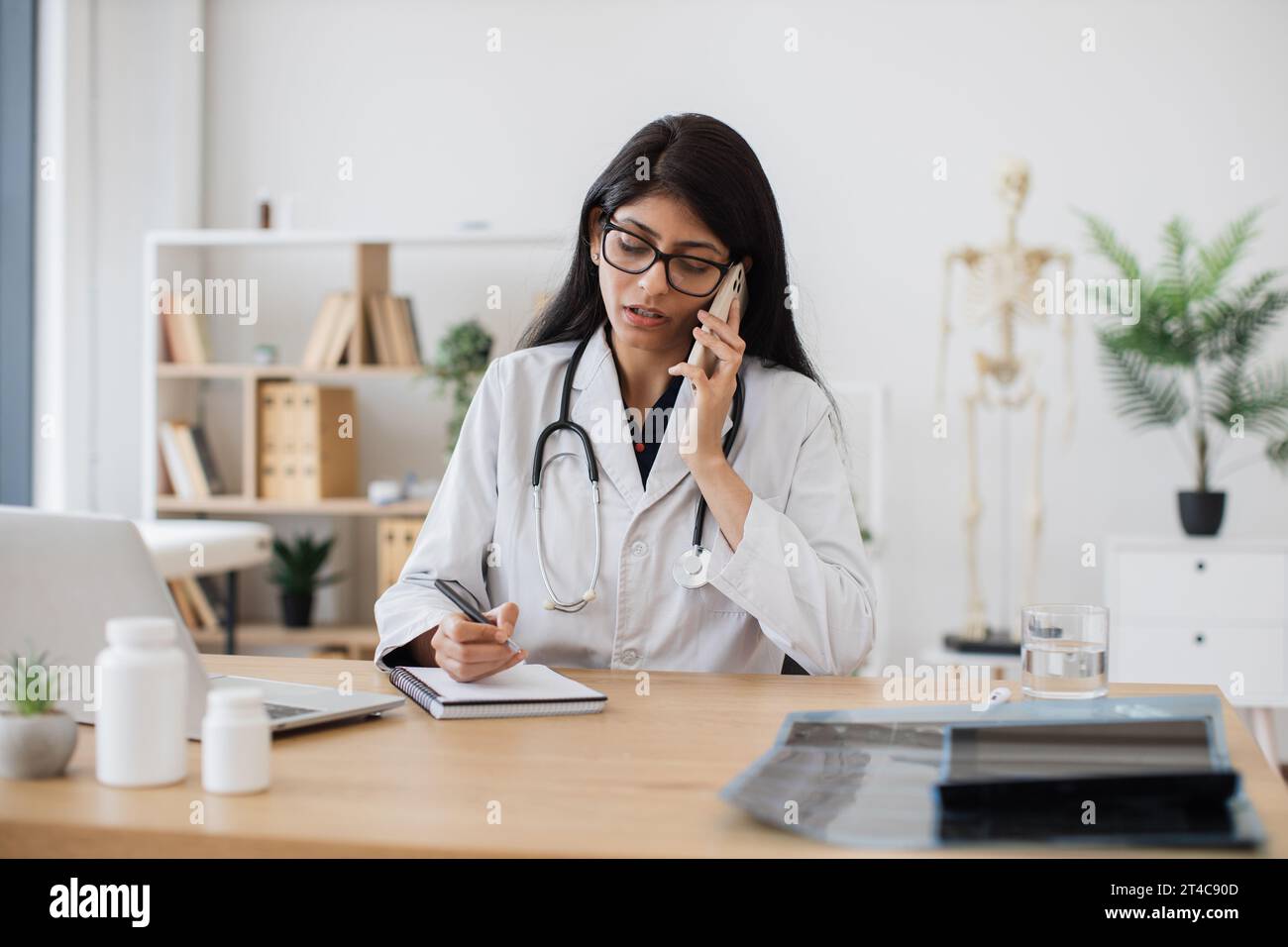 Experienced indian doctor talking on modern smartphone with ill patient while sitting at personal workplace in modern hospital. Mature female writing Stock Photo