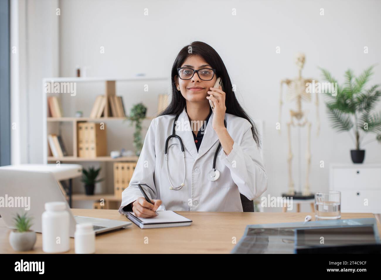Busy female doctor speaking on modern smartphone and writing patient problems in notebook. Focused indian woman with stethoscope on neck sitting at ta Stock Photo