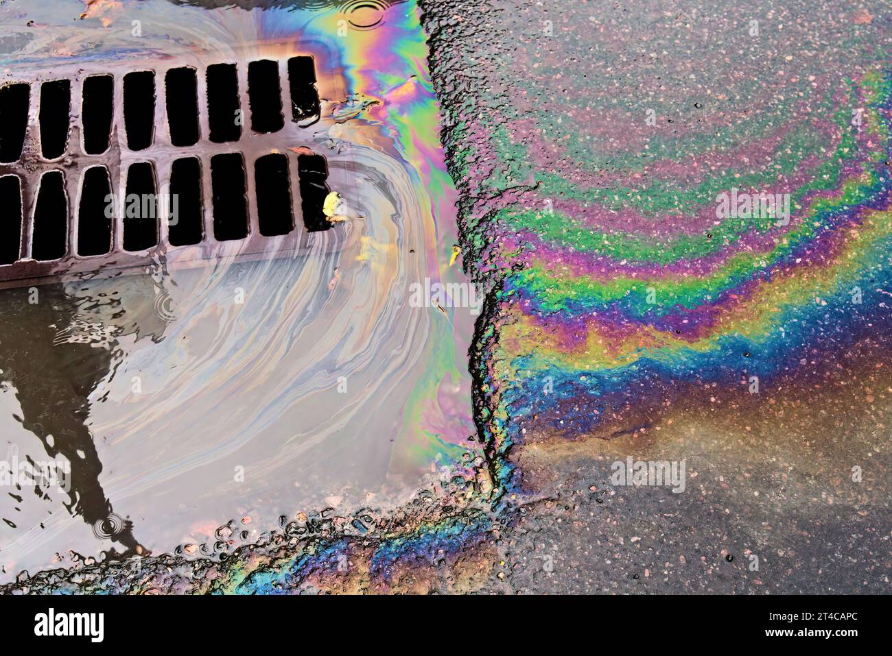 An oil slick against the backdrop of an asphalt road flows into a storm drain through a grate. Environmental problems of water pollution. Stock Photo