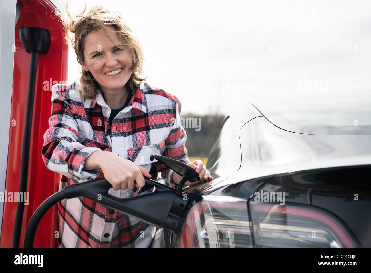 Woman in a plaid shirt charging an electric car. Stock Photo