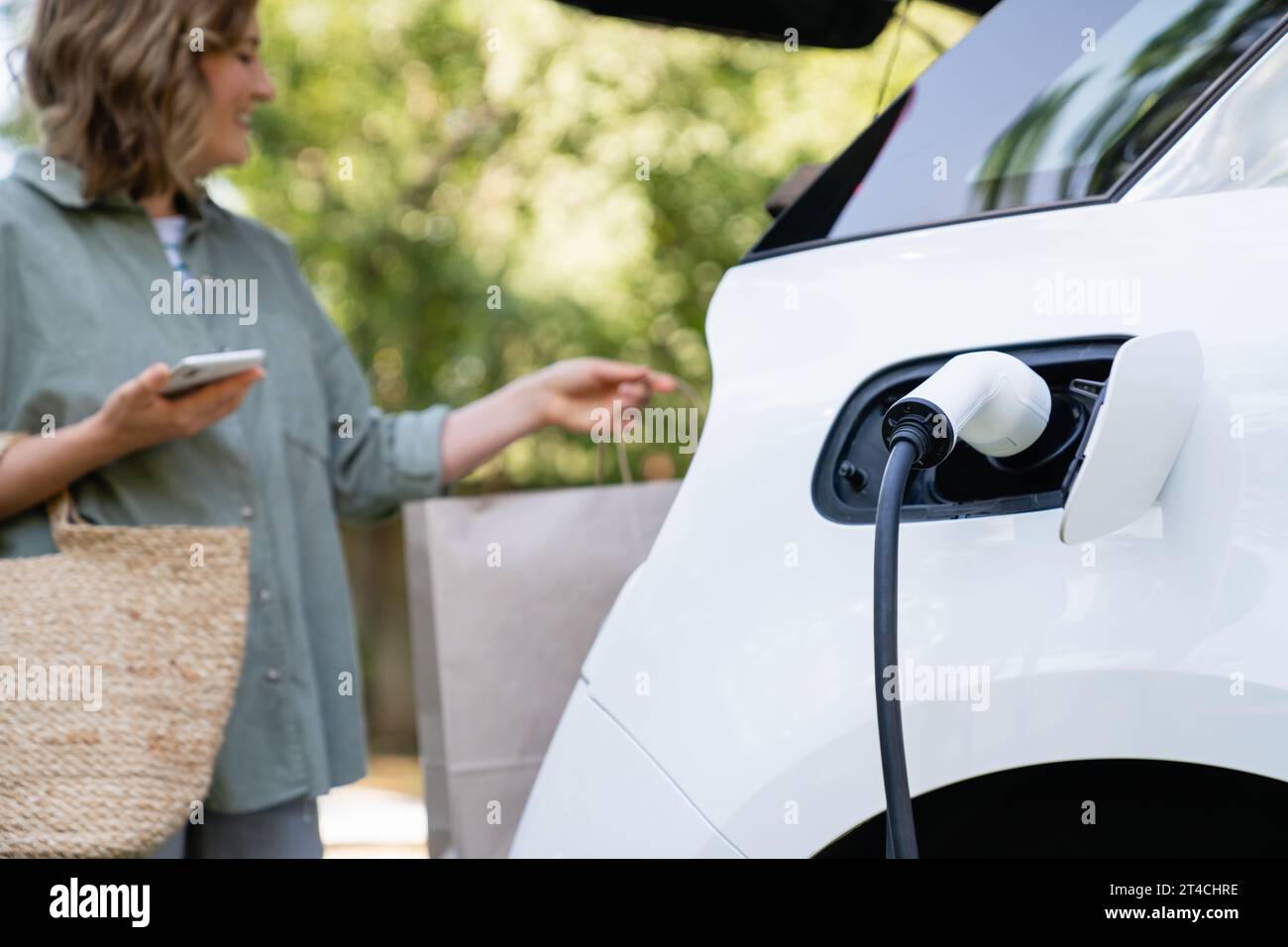 Woman takes a shopping bag from a trunk of charging electric car. Stock Photo