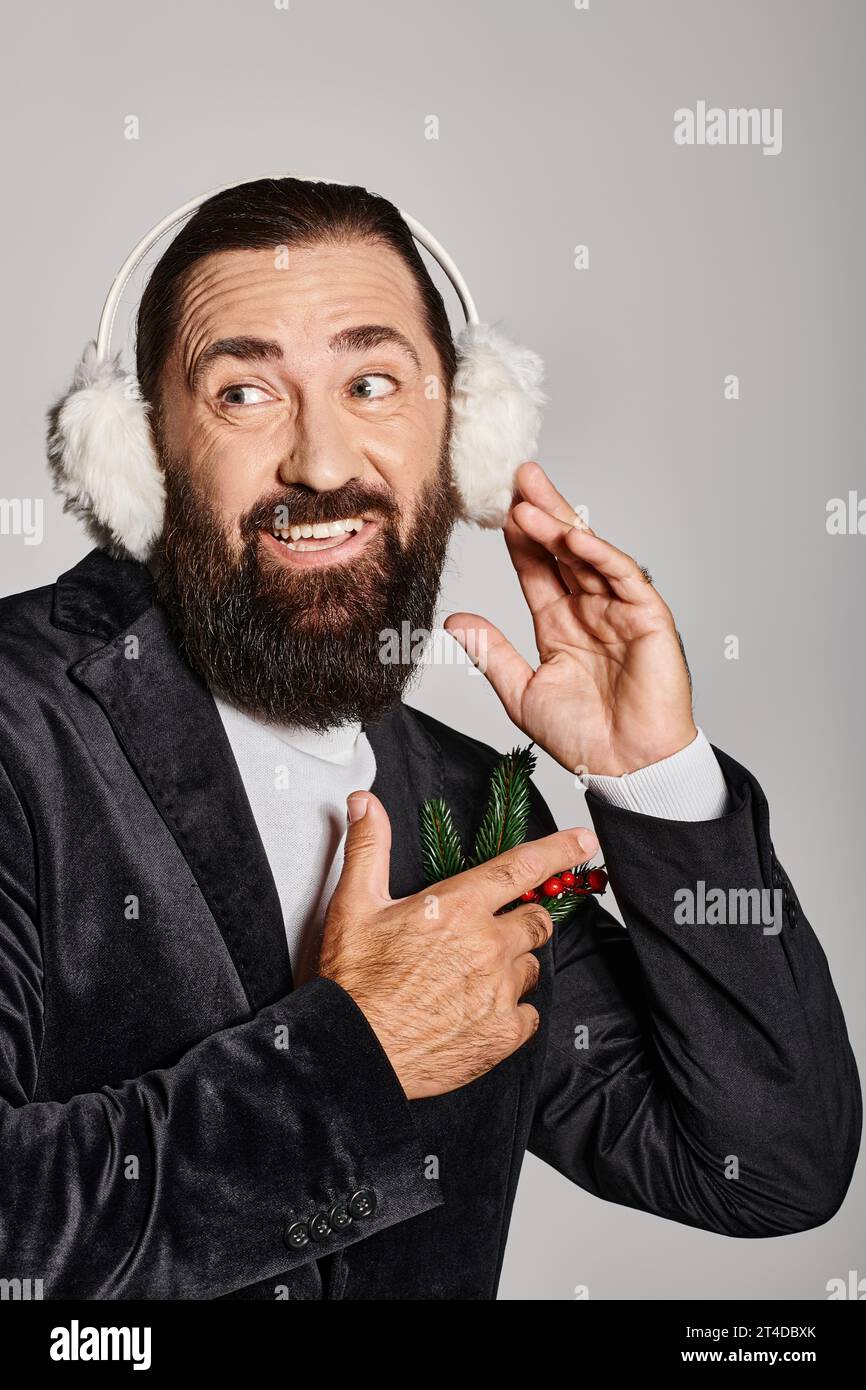 cheerful bearded man in suit with Christmas spruce in pocket wearing ear muffs on grey backdrop Stock Photo