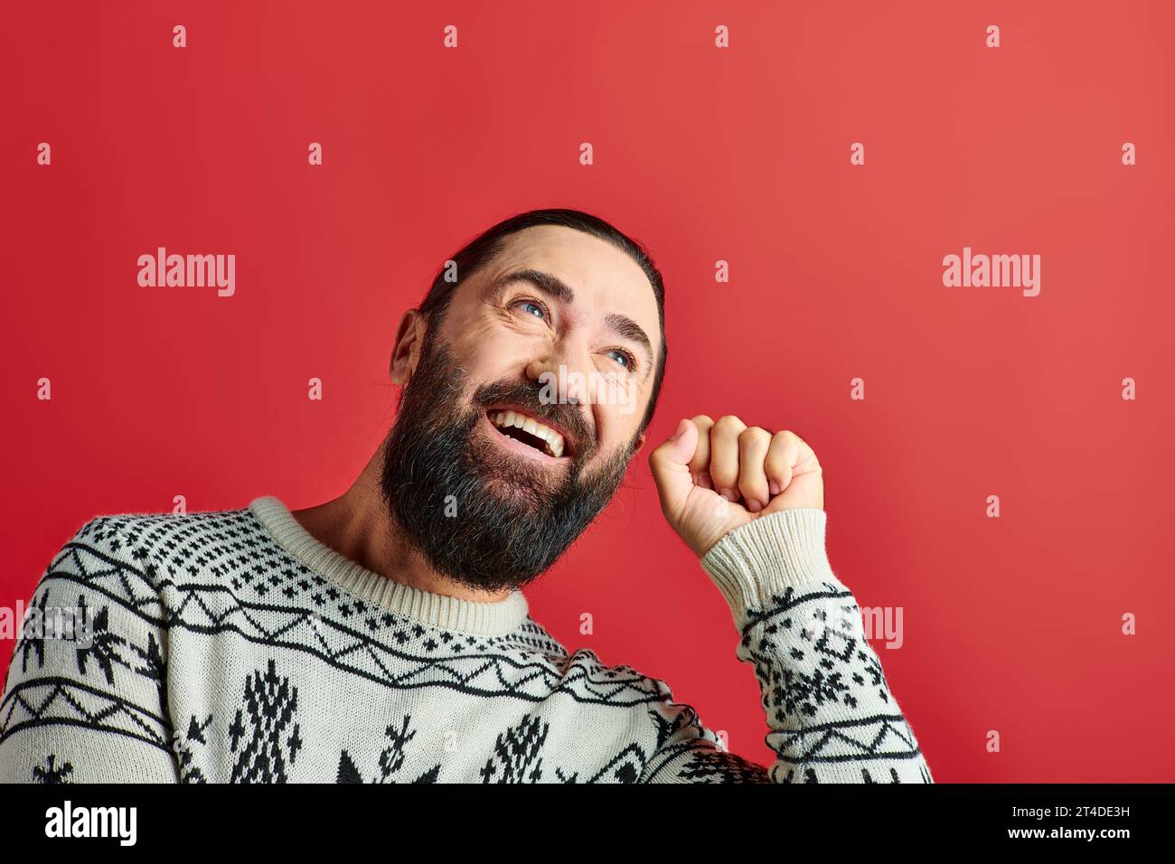 cheerful bearded man in winter sweater with ornament knocking on red backdrop, Merry Christmas Stock Photo