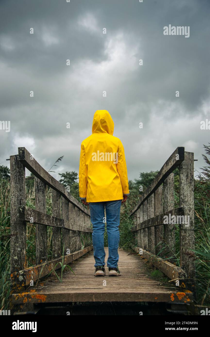 rear view of a woman wearing a yellow coat with hood standing on a wooden footbridge. Stock Photo