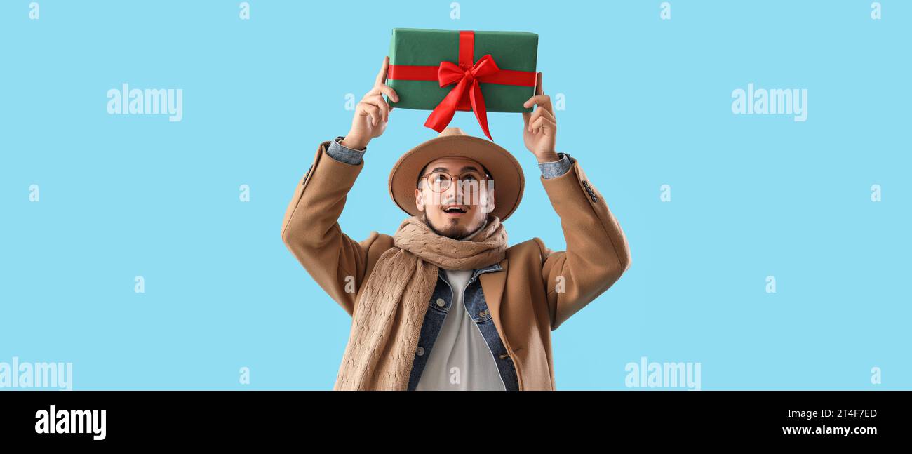 Stylish young man in winter clothes holding gift on light blue background Stock Photo
