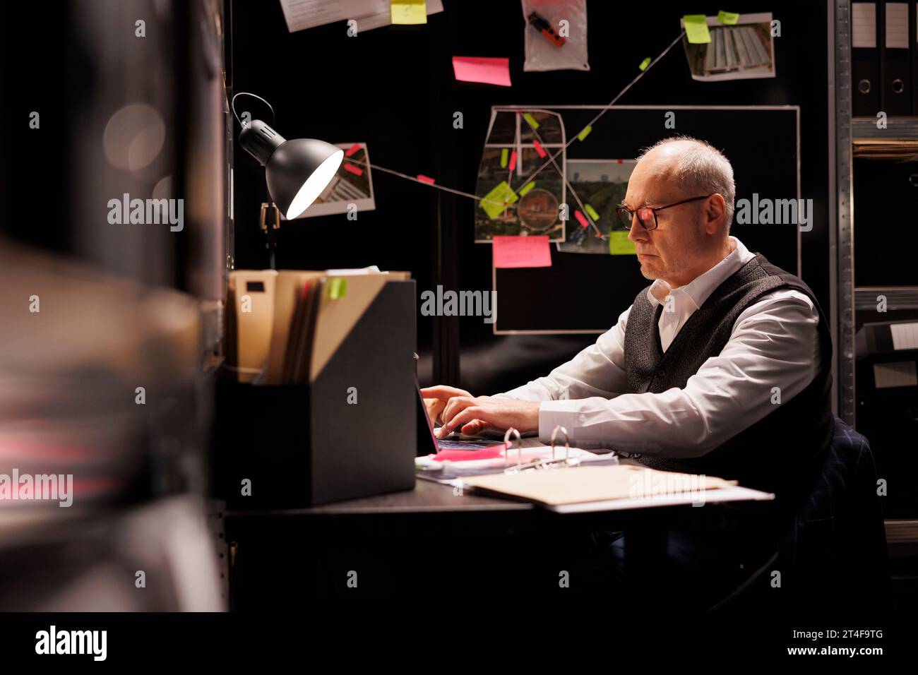 Elderly man inspector sitting at desk table in evidence room, working late at night at missing person case. Senior private detective analyzing victim files, looking at crime scene report Stock Photo