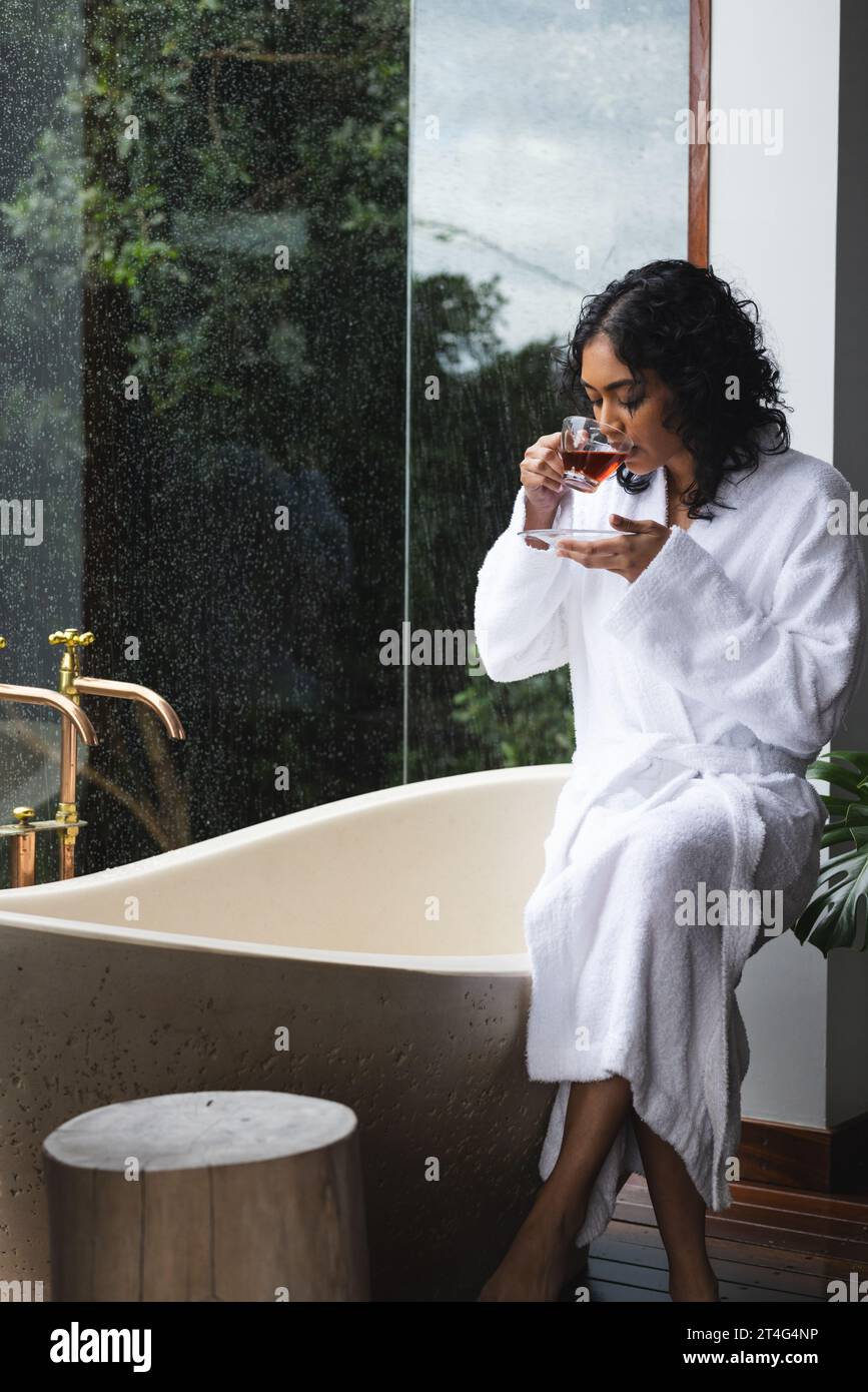 Happy biracial woman in bathrobe sitting on bath and drinking tea in sunny bathroom Stock Photo