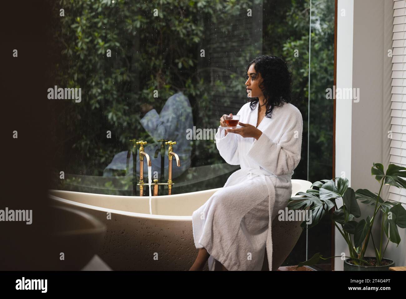 Happy biracial woman in bathrobe sitting on bath with tea and looking out window in sunny bathroom Stock Photo