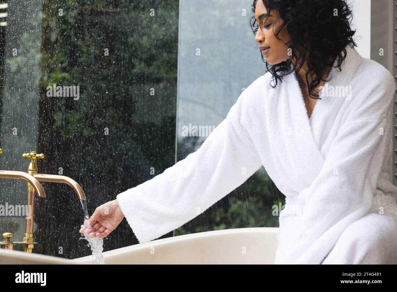 Happy biracial woman in bathrobe sitting on bath and checking water in sunny bathroom Stock Photo