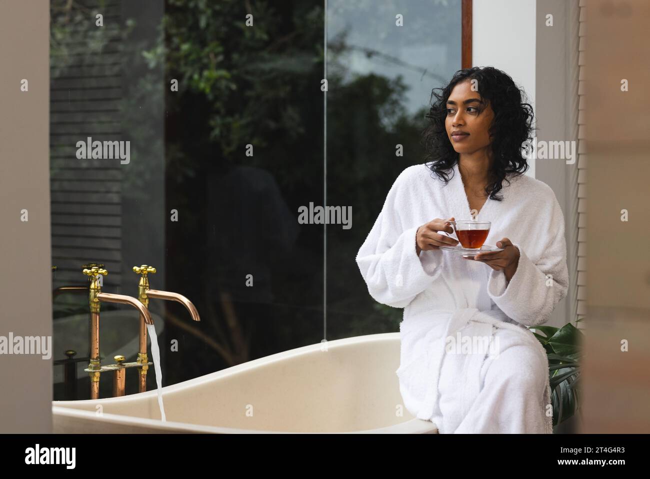 Happy biracial woman in bathrobe sitting on bath with tea and looking out window in sunny bathroom Stock Photo