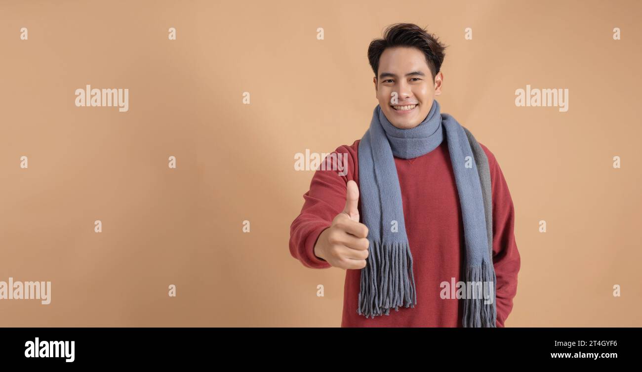 Happy and positive Asian man in red sweater and scarf giving thumbs up with ad empty space isolated on beige color background. Stock Photo