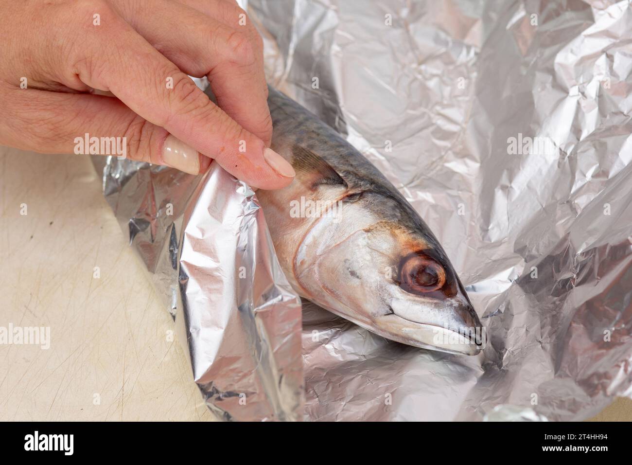 Woman hand wrapping in tinfoil whole raw mackerel fish before baking Stock Photo