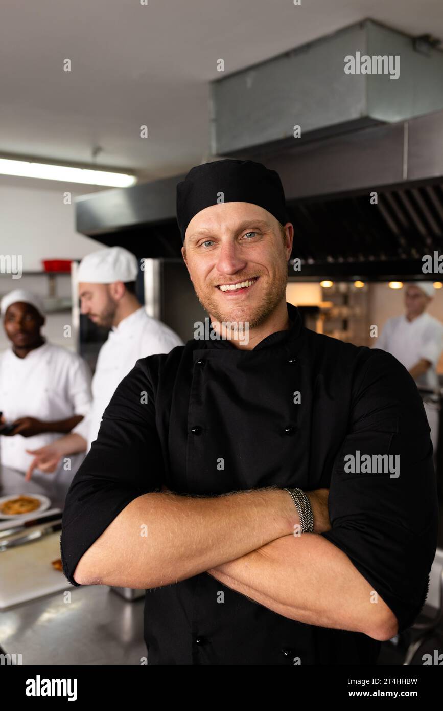 Diverse happy male head chef with arms crossed standing against students cooking in background Stock Photo