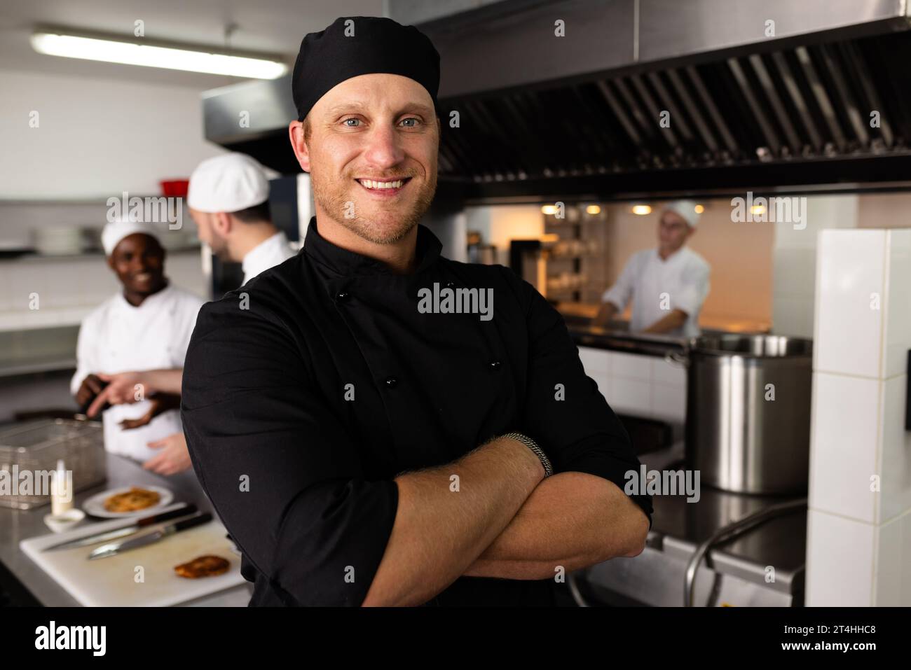 Diverse head chef with arms crossed standing against male students cooking in background Stock Photo
