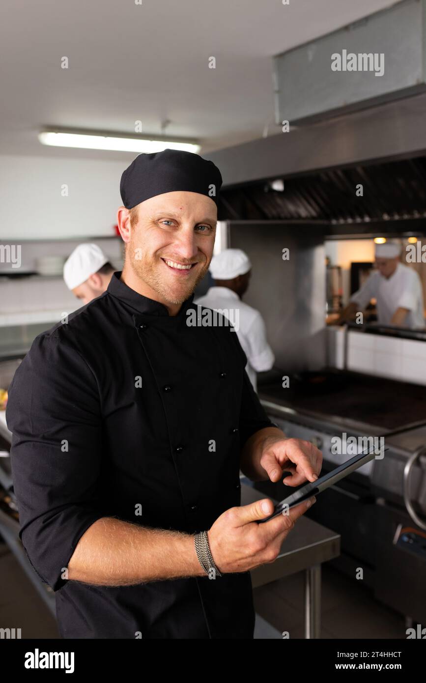 Diverse smiling chef teacher in black uniform using tablet while students working in background Stock Photo